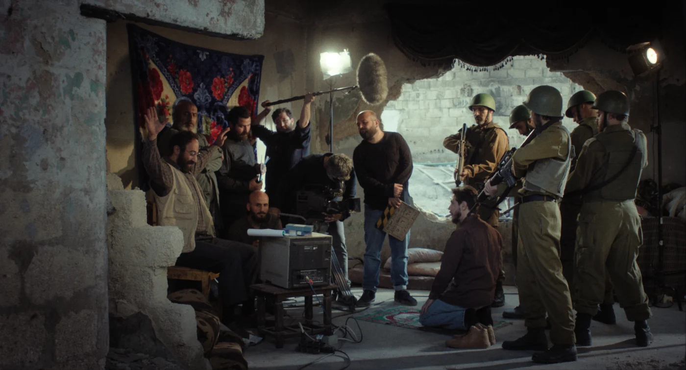 Mehrere Personen in Militäruniformen stehen in einem abgedunkelten Raum mit beschädigten Wänden. Einige tragen Helme und halten Gewehre. Weitere Personen tragen Alltagskleidung und bedienen Filmtechnik wie eine Tonangel, eine Filmklappe und Kameras. Im Hintergrund hängt ein gemustertes Tuch an der Wand, davor stehen elektronische Geräte auf einem Tisch. Sandsäcke sind im Hintergrund gestapelt. [erstellt mit KI]