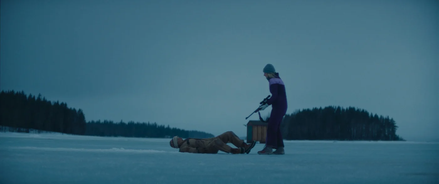 Eine Person in dunkler Winterkleidung steht vor einer am Boden liegenden Person. Die stehende Person trägt eine Mütze und hält Gewehr in den Händen. Im Hintergrund sind bewaldete Ufer und ein bewölkter Himmel zu sehen. [erstellt mit KI]