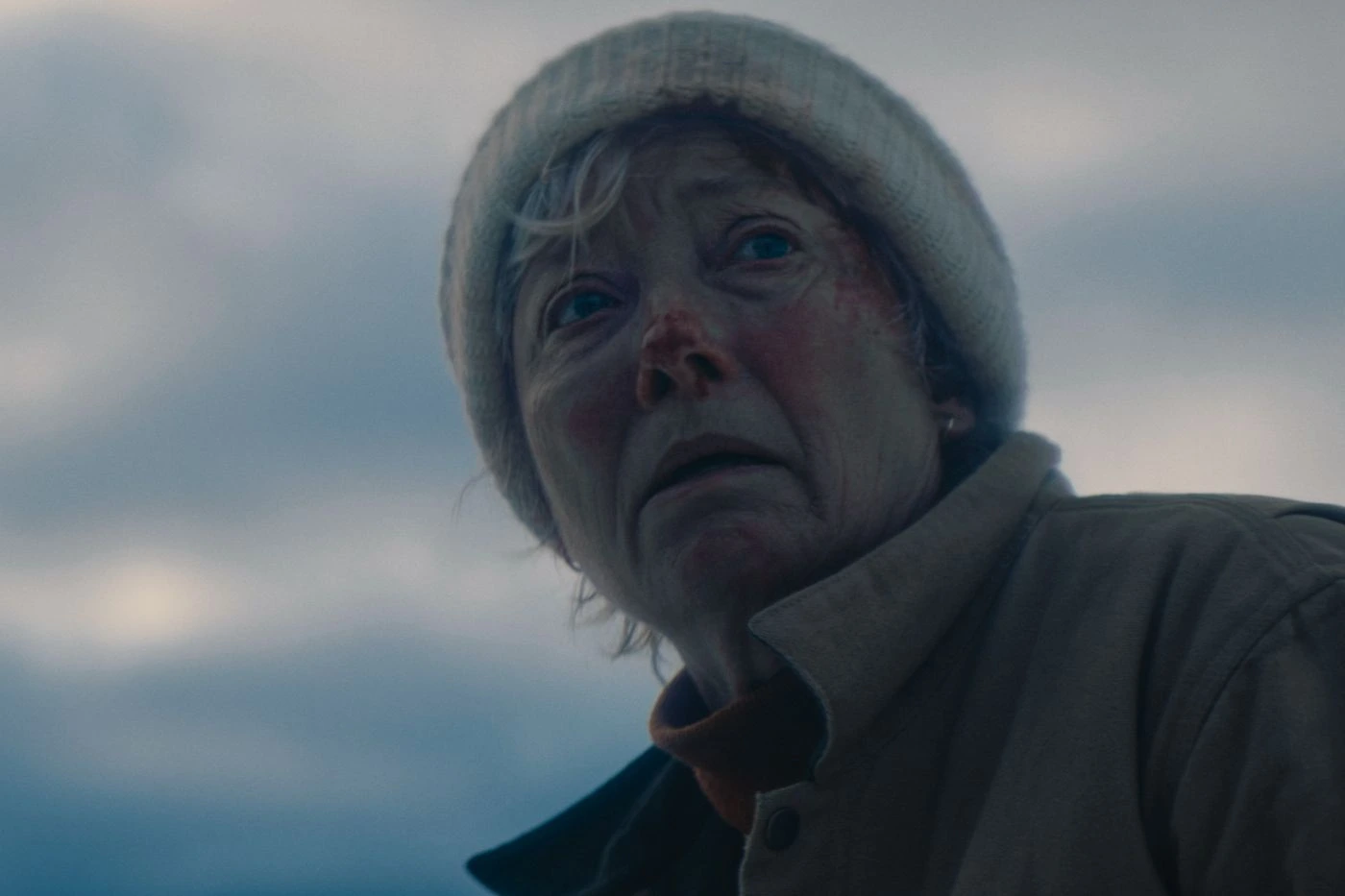 Eine Person trägt eine helle Strickmütze und eine Jacke mit Kragen. Der Hintergrund zeigt einen wolkenverhangenen Himmel mit gedämpften Farben. [erstellt mit KI]