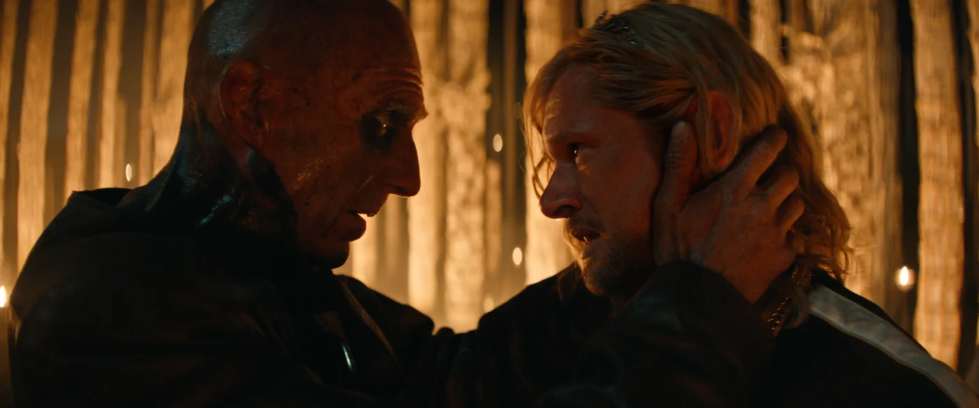 Zwei Personen stehen sich gegenüber, eine Person hält die andere am Hinterkopf. Beide tragen dunkle Kleidung. Im Hintergrund sind vertikale, beleuchtete Streifen zu sehen, die eine warme Lichtstimmung erzeugen. [erstellt mit KI]