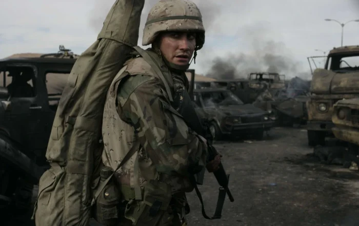 Eine Person in Tarnuniform und Helm steht zwischen ausgebrannten Fahrzeugwracks auf einer zerstörten Straße. Die Person trägt einen großen, länglichen Rucksack auf dem Rücken. Im Hintergrund sind mehrere zerstörte Fahrzeuge und aufsteigende Rauchwolken zu sehen. [erstellt mit KI]