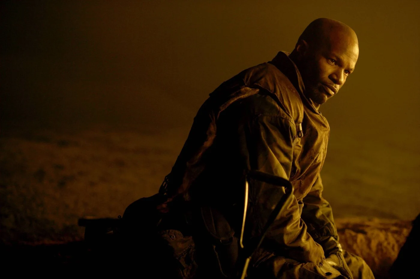 Eine Person mit kahlrasiertem Kopf sitzt in einem sandfarbenen, militärisch wirkenden Overall auf dem Boden. Die Szene ist in warmes, gelbliches Licht getaucht. Im Vordergrund ist ein Gewehr mit markantem Schaft zu erkennen, das an die Schulter der Person gelehnt ist. [erstellt mit KI]