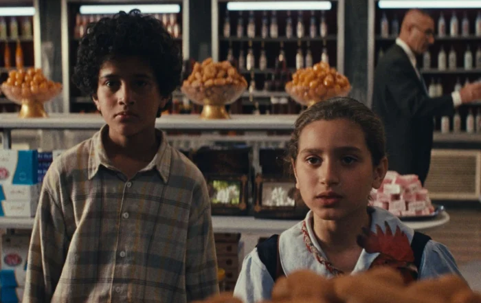 Zwei Kinder stehen nebeneinander in einem Lebensmittelgeschäft. Im Hintergrund sind Regale mit Flaschen und Körben voller Backwaren zu sehen. Ein Erwachsener im Anzug bewegt sich durch den Laden. Die Kinder tragen helle, gemusterte Kleidung und stehen vor einem Tisch mit Gebäck. [erstellt mit KI]