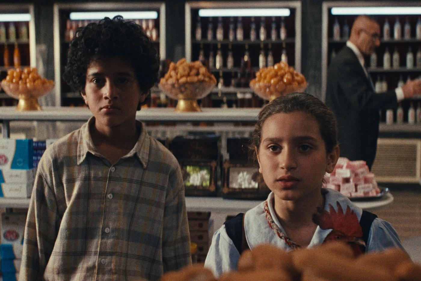 Zwei Kinder stehen nebeneinander in einem Lebensmittelgeschäft. Im Hintergrund sind Regale mit Flaschen und Körben voller Backwaren zu sehen. Ein Erwachsener im Anzug bewegt sich durch den Laden. Die Kinder tragen helle, gemusterte Kleidung und stehen vor einem Tisch mit Gebäck. [erstellt mit KI]