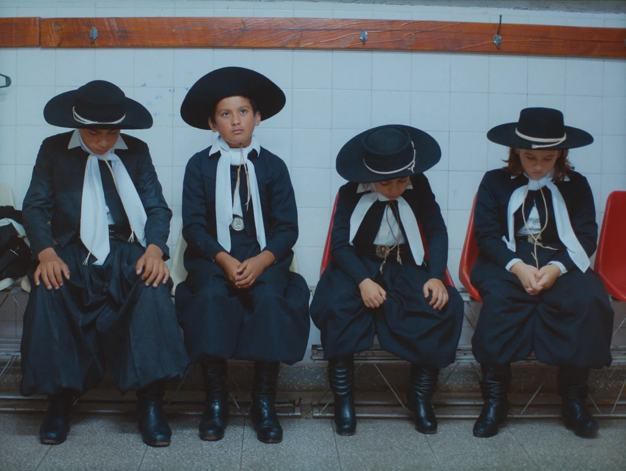 Vier Personen sitzen nebeneinander auf roten Kunststoffstühlen vor einer weiß gefliesten Wand. Sie tragen dunkle, traditionelle Kleidung mit weiten Röcken oder Hosen, weißen Halstüchern und breiten, flachen Hüten. Die Hände liegen auf dem Schoß, die Beine sind geschlossen. Die Schuhe sind ebenfalls dunkel und glänzend. Über den Personen verläuft eine Holzleiste an der Wand. [erstellt mit KI]
