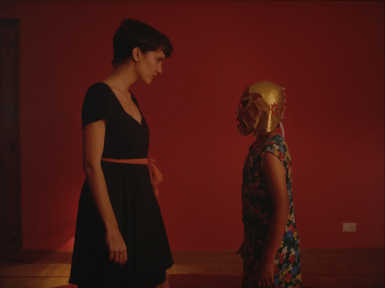 Eine Person in einem schwarzen Kleid mit orangefarbenem Gürtel steht in einem Raum mit roten Wänden. Gegenüber steht ein Kind mit goldener Maske und bunter Kleidung. Beide Personen stehen sich frontal gegenüber. Der Raum ist spärlich beleuchtet und wirkt leer, bis auf eine sichtbare Steckdose an der Wand. [erstellt mit KI]