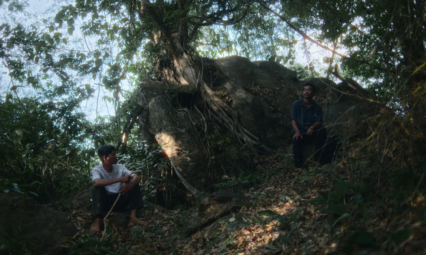 Zwei Personen sitzen in einem bewaldeten, schattigen Gelände auf dem Boden und auf einem Felsen. Im Hintergrund steht ein großer Baum mit ausladenden Wurzeln, die sich über einen Felsen ziehen. Sonnenlicht fällt durch das dichte Blätterdach und erzeugt helle Flecken auf dem Waldboden. [erstellt mit KI]
