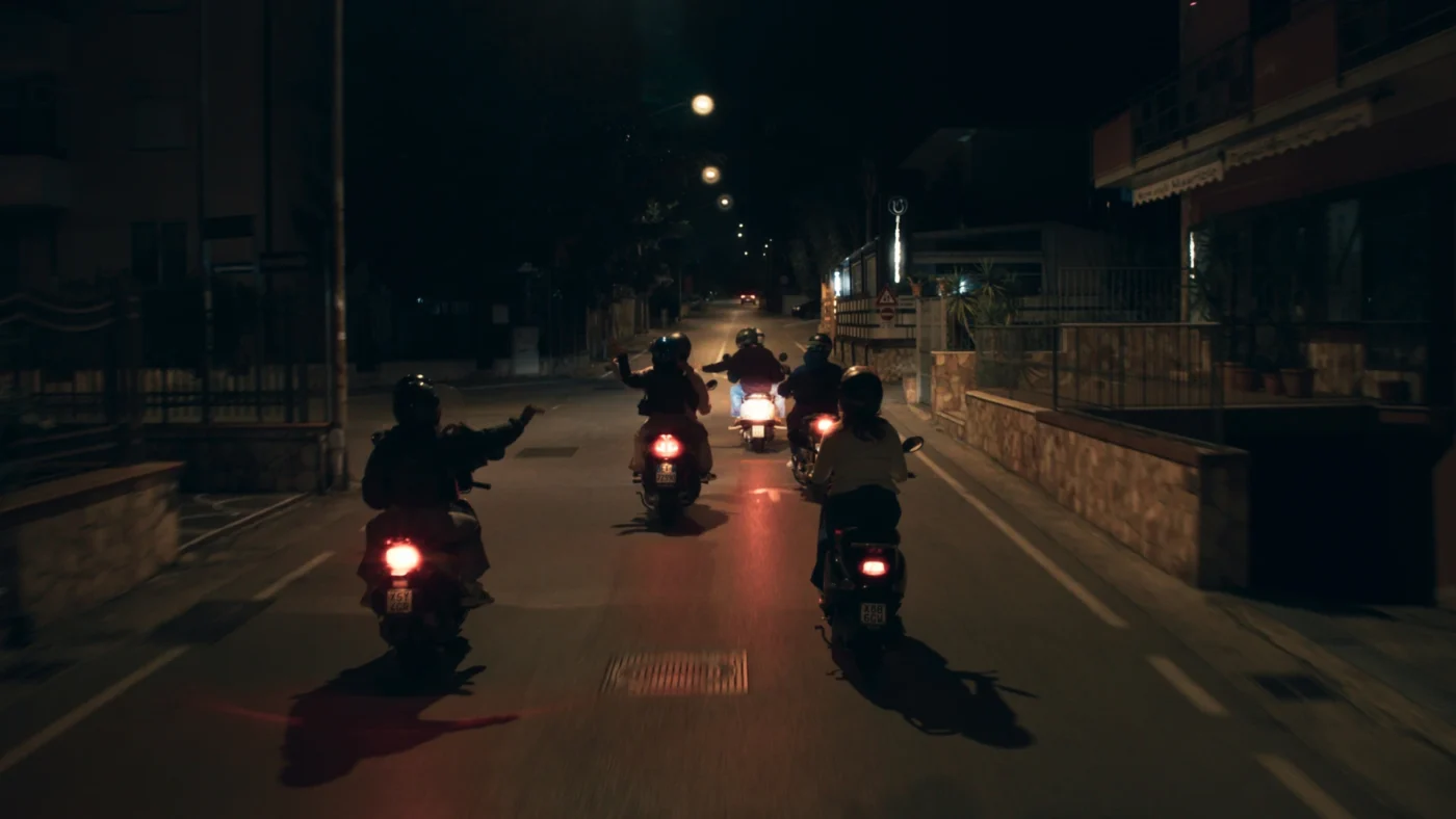 Nachts fahren mehrere Personen mit Motorrollern eine beleuchtete Straße entlang. Sie sind von hinten zu sehen, tragen Helme und fahren in einer Gruppe. Die Straße ist von Häusern gesäumt, Straßenlaternen erhellen die Szene. [erstellt mit KI]