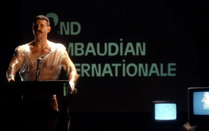 Szenenbild Uchronia: Eine Person steht an einem Rednerpult, trägt ein transparentes Oberteil und wird von einer starken Lichtquelle beleuchtet. Im Hintergrund ist eine große Projektion mit weißen Buchstaben auf schwarzem Grund zu sehen. Rechts im Bild steht ein Röhrenfernseher, auf dessen Bildschirm ein Gesicht und ein statisches Bild zu erkennen sind. [erstellt mit KI]