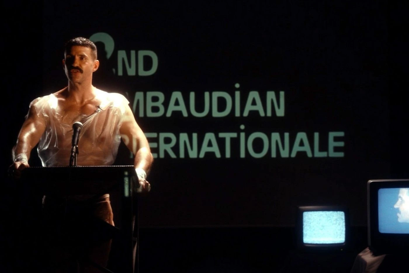 Szenenbild Uchronia: Eine Person steht an einem Rednerpult, trägt ein transparentes Oberteil und wird von einer starken Lichtquelle beleuchtet. Im Hintergrund ist eine große Projektion mit weißen Buchstaben auf schwarzem Grund zu sehen. Rechts im Bild steht ein Röhrenfernseher, auf dessen Bildschirm ein Gesicht und ein statisches Bild zu erkennen sind. [erstellt mit KI]