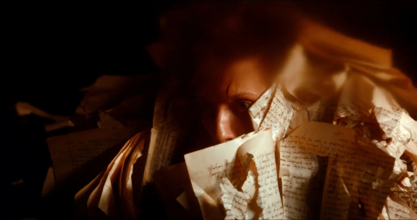 Zerrissene Papierblätter mit handgeschriebenem Text liegen in chaotischer Anordnung. Die Schrift ist dunkel und kontrastreich, einzelne Wörter und Zeilen sind deutlich erkennbar. Warmes Licht fällt auf die Szene und erzeugt starke Schatten. [erstellt mit KI]