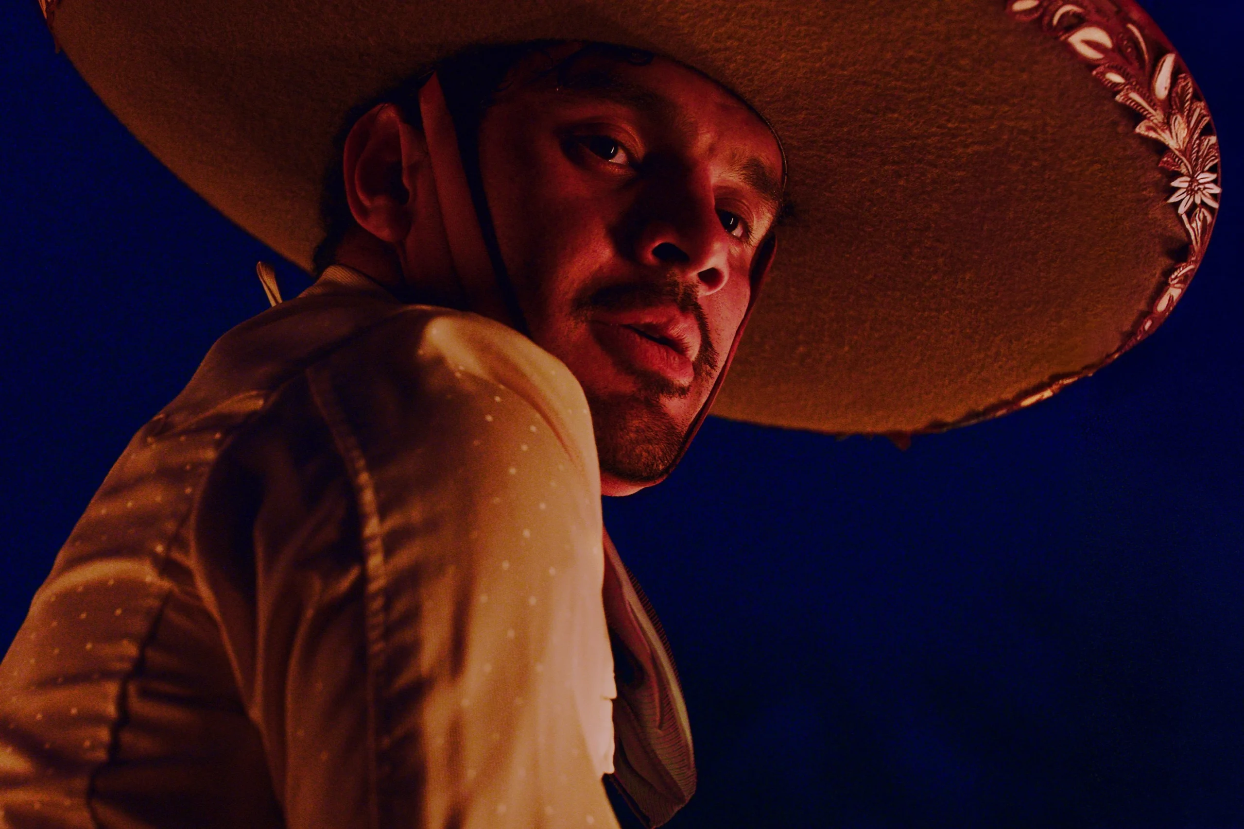 Ein Mensch trägt einen großen, runden Hut mit dekorativem Muster am Rand. Das Hemd ist hell und mit feinen Punkten versehen. Der Hintergrund ist dunkelblau und erzeugt einen starken Kontrast zu der Kleidung und dem Hut. [erstellt mit KI]