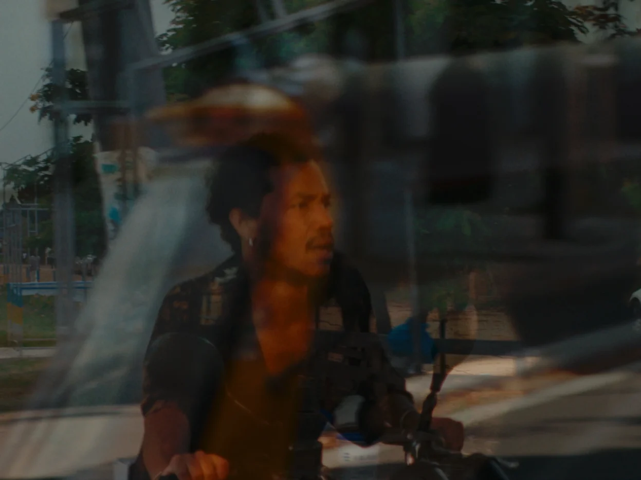 Motorradfahrer mit dunkler Kleidung fährt auf einer Straße, im Vordergrund ist ein verschwommenes Fahrzeug zu sehen. Im Hintergrund sind Bäume und ein Zaun erkennbar. [erstellt mit KI]