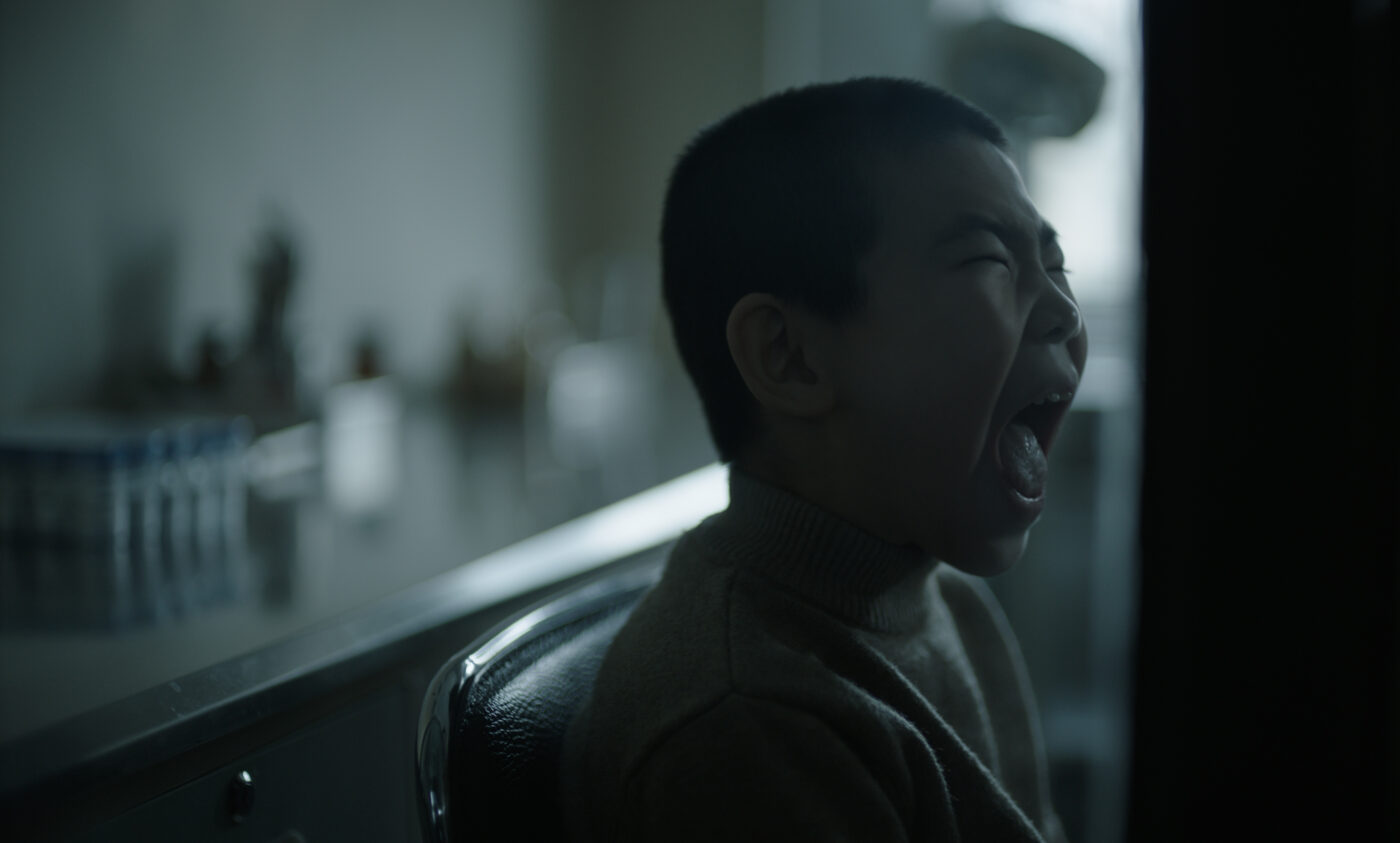 Eine Person sitzt auf einem dunklen Stuhl in einem schwach beleuchteten Raum. Im Hintergrund sind unscharfe Gegenstände auf einer Oberfläche zu erkennen. Die Lichtstimmung ist gedämpft und kühl. [erstellt mit KI]