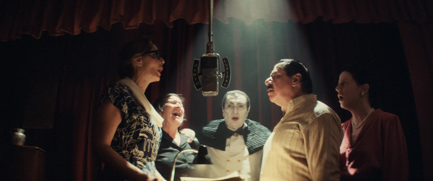 Mehrere Personen stehen eng beieinander unter einem großen, zentral hängenden Mikrofon. Sie tragen formelle oder halbformelle Kleidung, darunter ein gemustertes Kleid, ein Hemd und ein Sakko mit Fliege. Im Hintergrund ist ein schwerer, dunkelroter Vorhang zu sehen, durch den ein Lichtstrahl auf die Gruppe fällt. [erstellt mit KI]