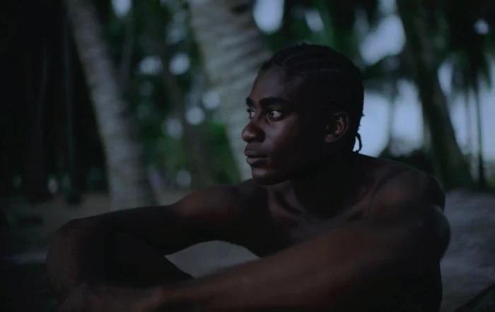 Szenenbild des Films Paradise: Eine Person mit geflochtenen Haaren sitzt im Halbdunkel unter Palmen. Die Arme sind locker um die Knie gelegt. Im Hintergrund sind unscharfe Palmenstämme und diffuse Lichtverhältnisse zu erkennen. [erstellt mit KI]