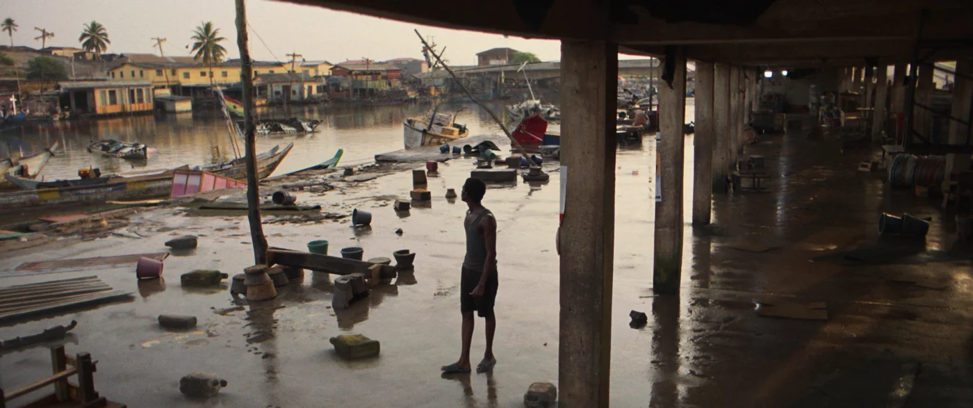 Ein überdachter, offener Bereich mit Betonboden, auf dem verstreut Gegenstände wie Eimer, Kisten und Steine liegen. Im Hintergrund sind ein Fluss oder Hafenbecken, mehrere bunte Boote und Häuser zu sehen. Eine Person steht im Halbschatten und blickt auf den Boden. Das Licht ist gedämpft, die Szene wirkt ruhig und verlassen.