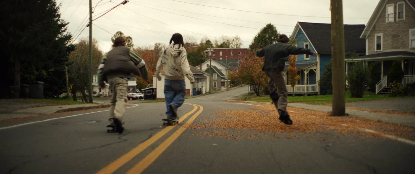Drei Jugendliche fahren auf Skateboards eine asphaltierte Straße entlang. Die Szene zeigt eine herbstliche Wohngegend mit Laub auf der Straße, Bäumen und Häusern im Hintergrund. Die Jugendlichen tragen Jacken und lange Hosen. Die Perspektive ist leicht von hinten, sodass die Gesichter nicht zu erkennen sind.