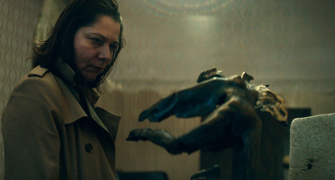 Eine Person mit langen, dunklen Haaren trägt einen beigefarbenen Mantel und steht in einem Raum mit gemusterter Tapete. Im Vordergrund befindet sich eine große, dunkle Skulptur in Form einer Hand mit ausgestreckten Fingern. [erstellt mit KI]