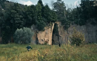 Szenenbild aus "Mambo Kids": Zu sehen ist eine Wiese mit hohem Gras in einer natürlichen Landschaft. Zwei Personen in dunkler Kleidung bewegen sich durch das Gras auf eine große Felswand zu. In der Mitte der Felswand befindet sich eine schmale, hohe Öffnung wie ein natürlicher Durchgang. Die Felswand ist von Bäumen und Sträuchern umgeben, darüber ein heller, leicht bewölkter Himmel. [erstellt mit KI]