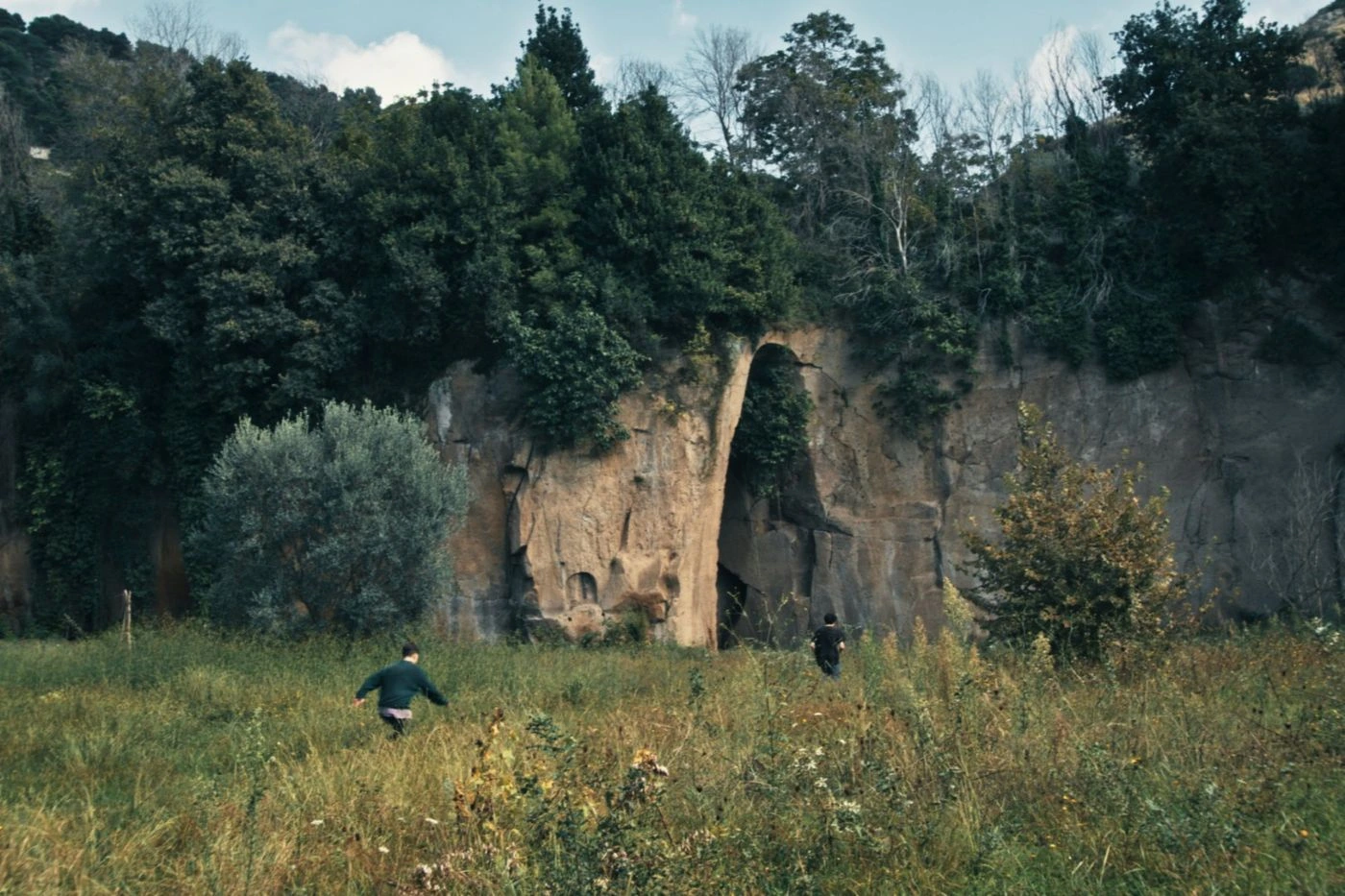 Szenenbild aus "Mambo Kids": Zu sehen ist eine Wiese mit hohem Gras in einer natürlichen Landschaft. Zwei Personen in dunkler Kleidung bewegen sich durch das Gras auf eine große Felswand zu. In der Mitte der Felswand befindet sich eine schmale, hohe Öffnung wie ein natürlicher Durchgang. Die Felswand ist von Bäumen und Sträuchern umgeben, darüber ein heller, leicht bewölkter Himmel. [erstellt mit KI]