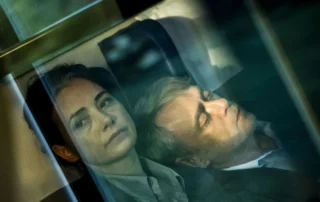 Szenenbild aus Gelbe Briefe: Zwei Personen sitzen nebeneinander in einem Zugabteil. Die Szene ist durch eine Glasscheibe aufgenommen, wodurch Spiegelungen und Lichtreflexe sichtbar sind. Beide tragen helle Oberteile und lehnen sich entspannt zurück. Der Hintergrund zeigt die Kopfstützen der Sitze und einen Teil des Zugfensters. [erstellt mit KI]