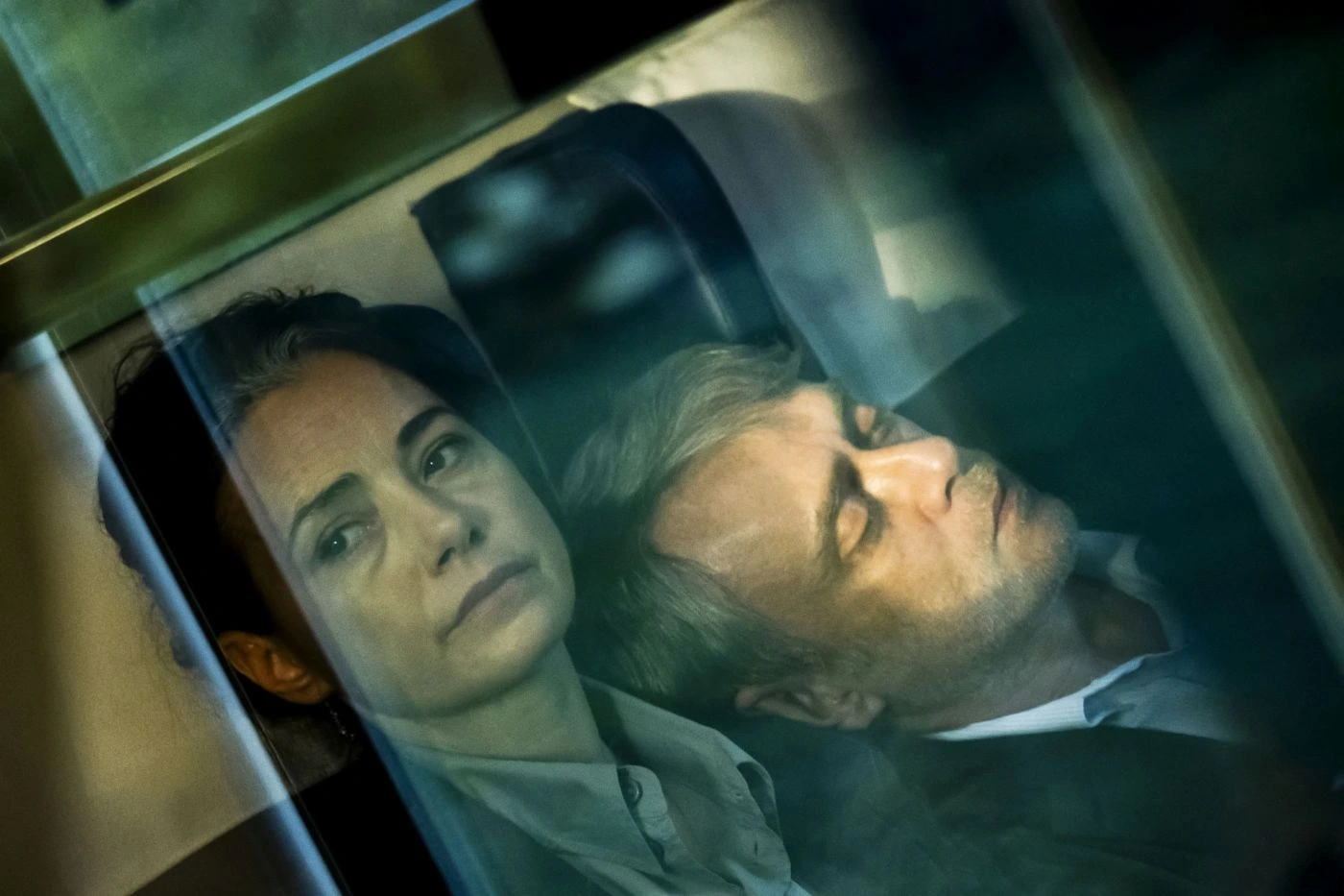 Szenenbild aus Gelbe Briefe: Zwei Personen sitzen nebeneinander in einem Zugabteil. Die Szene ist durch eine Glasscheibe aufgenommen, wodurch Spiegelungen und Lichtreflexe sichtbar sind. Beide tragen helle Oberteile und lehnen sich entspannt zurück. Der Hintergrund zeigt die Kopfstützen der Sitze und einen Teil des Zugfensters. [erstellt mit KI]