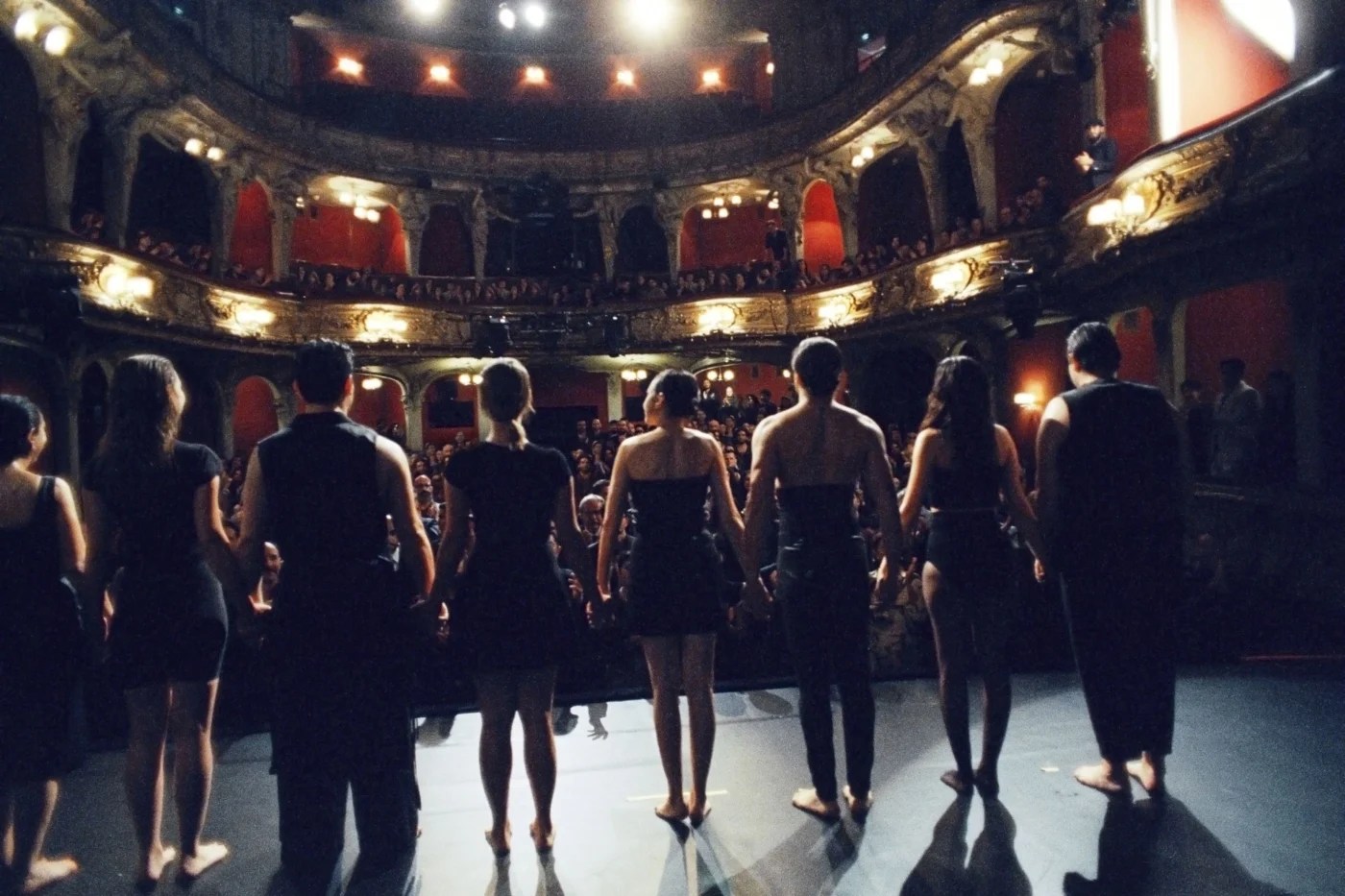 Eine Gruppe von Menschen steht barfuß auf einer Bühne und blickt in Richtung Publikum. Die Personen tragen dunkle Kleidung und halten sich an den Händen. Im Hintergrund sind die Ränge eines Theaters mit Zuschauern zu sehen, die von warmem Licht beleuchtet werden. Die Architektur zeigt verzierte Balustraden und rote Wände. [erstellt mit KI]