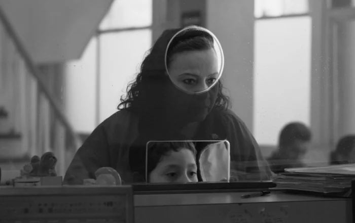 Szenenbild aus Moscas: Eine Frau wartet vor einem Tisch mit großer Glasscheibe und schaut durch ein Loch in der Scheibe. Ein kleiner Junge steht vor ihr und lugt durch ein weiteres Loch in der Scheibe.