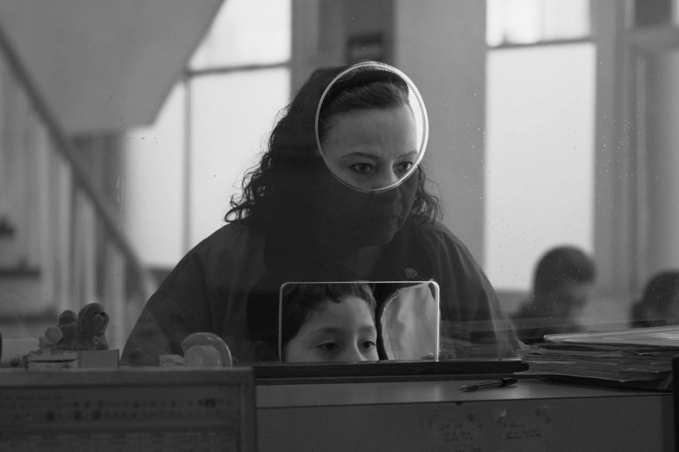 Szenenbild aus Moscas: Eine Frau wartet vor einem Tisch mit großer Glasscheibe und schaut durch ein Loch in der Scheibe. Ein kleiner Junge steht vor ihr und lugt durch ein weiteres Loch in der Scheibe.
