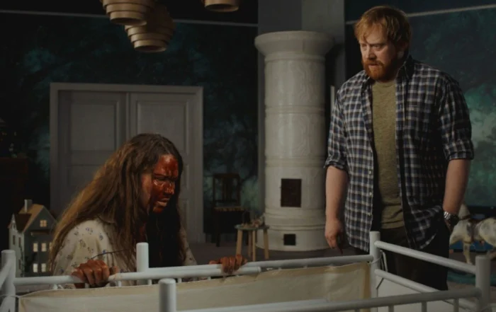 Szenenbild des Films "Nightborn": Ein Raum mit dunklen Wänden und einem weißen Kachelofen im Hintergrund. Im Vordergrund steht ein weißes Gitterbett, an dem eine Person mit langen, zerzausten Haaren festhält. Daneben steht eine weitere Person in kariertem Hemd und dunkler Hose. Im Hintergrund sind ein Puppenhaus und weitere Möbelstücke zu sehen. Die Beleuchtung ist gedämpft. [erstellt mit KI]