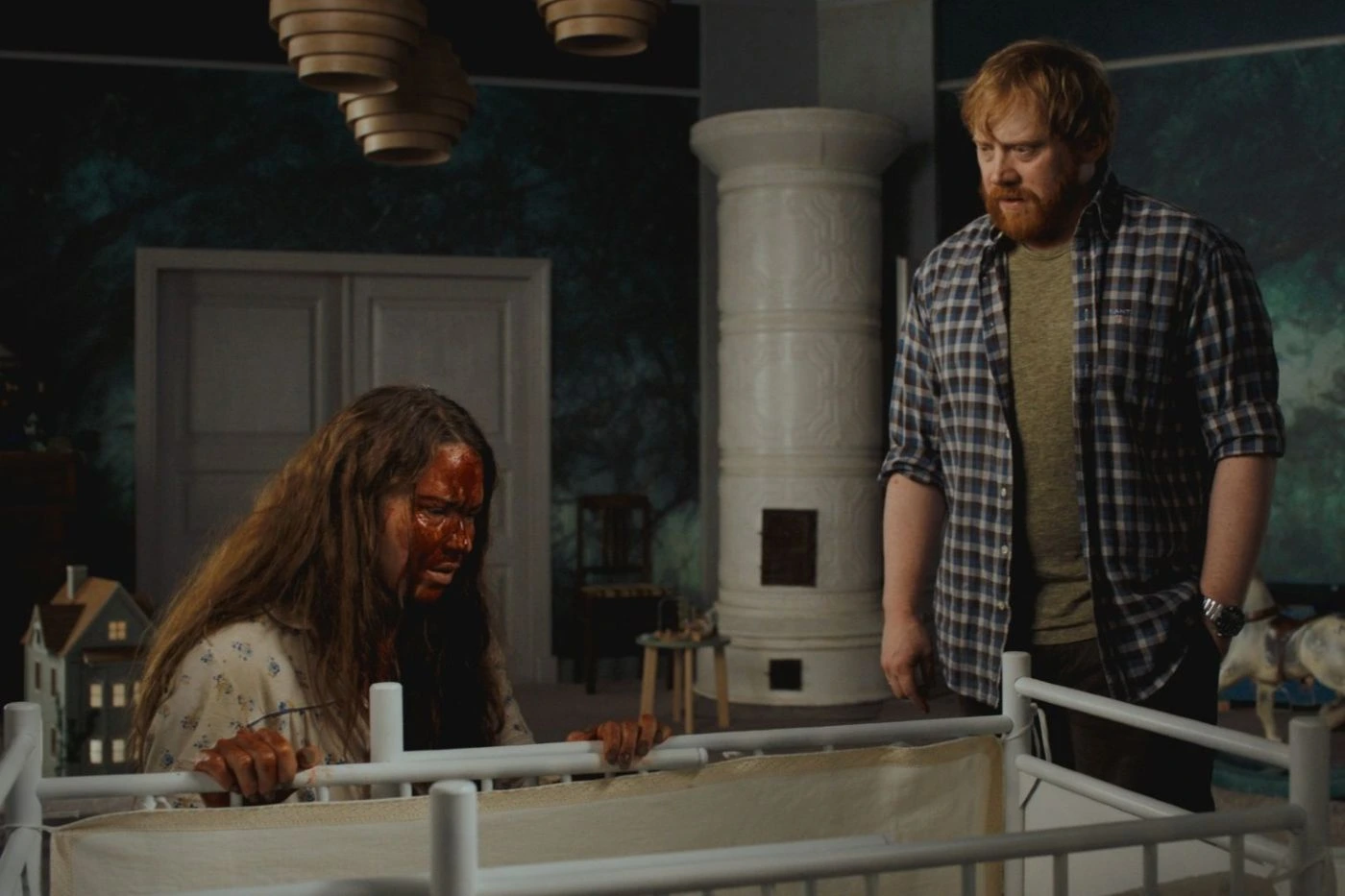 Szenenbild des Films "Nightborn": Ein Raum mit dunklen Wänden und einem weißen Kachelofen im Hintergrund. Im Vordergrund steht ein weißes Gitterbett, an dem eine Person mit langen, zerzausten Haaren festhält. Daneben steht eine weitere Person in kariertem Hemd und dunkler Hose. Im Hintergrund sind ein Puppenhaus und weitere Möbelstücke zu sehen. Die Beleuchtung ist gedämpft. [erstellt mit KI]