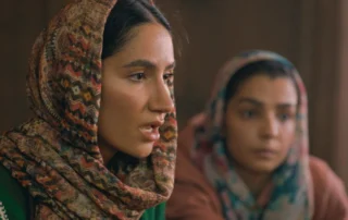 Zwei Personen sitzen nebeneinander in einem Innenraum. Beide tragen gemusterte Kopftücher, eines mit geometrischen Formen in verschiedenen Farben, das andere in gedeckten Tönen. Die Kleidung ist langärmelig und bedeckt die Arme. Der Hintergrund ist unscharf und zeigt warme Brauntöne. [erstellt mit KI]