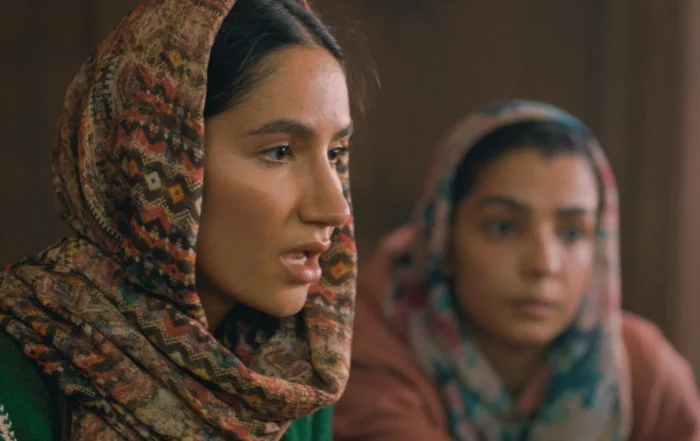 Zwei Personen sitzen nebeneinander in einem Innenraum. Beide tragen gemusterte Kopftücher, eines mit geometrischen Formen in verschiedenen Farben, das andere in gedeckten Tönen. Die Kleidung ist langärmelig und bedeckt die Arme. Der Hintergrund ist unscharf und zeigt warme Brauntöne. [erstellt mit KI]