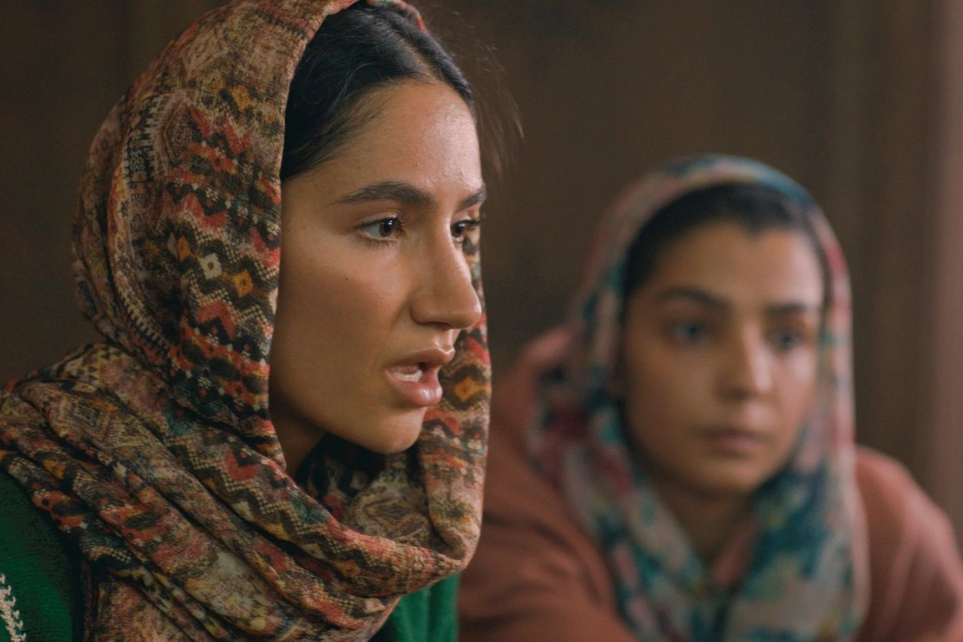 Zwei Personen sitzen nebeneinander in einem Innenraum. Beide tragen gemusterte Kopftücher, eines mit geometrischen Formen in verschiedenen Farben, das andere in gedeckten Tönen. Die Kleidung ist langärmelig und bedeckt die Arme. Der Hintergrund ist unscharf und zeigt warme Brauntöne. [erstellt mit KI]