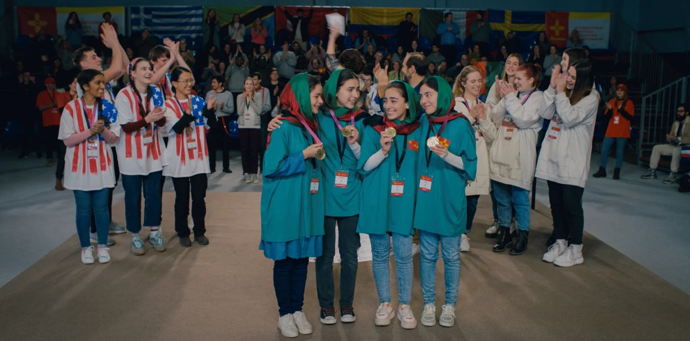 Mehrere Kinder stehen in der Mitte einer Halle und tragen türkisfarbene Umhänge mit roten Halstüchern. Sie halten Medaillen und kleine Gegenstände in den Händen. Im Hintergrund sind weitere Personen zu sehen, einige tragen weiße Jacken mit roten Streifen und halten blaue Objekte. Dahinter hängen verschiedene Landesflaggen an der Wand. [erstellt mit KI]