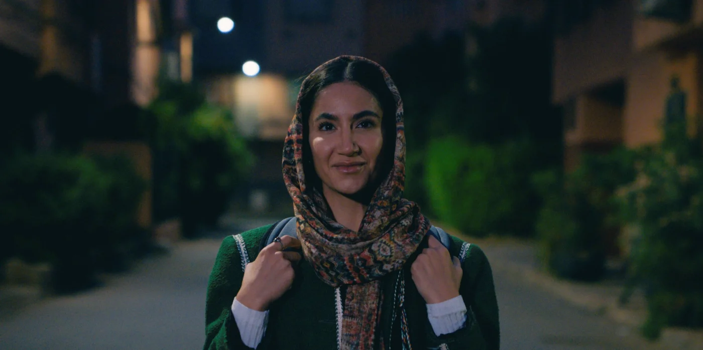 Eine Person steht auf einer nächtlichen Straße zwischen Gebäuden und hält die Träger eines Rucksacks mit beiden Händen. Die Person trägt ein gemustertes Kopftuch und einen dunklen Mantel mit hellen Streifen. Im Hintergrund sind unscharfe Lichter und grüne Pflanzen zu erkennen. [erstellt mit KI]