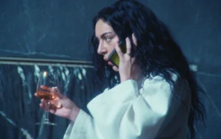 Szenenbild zum Film The Moment: Eine Person mit langen, dunklen, lockigen Haaren trägt einen hellen Bademantel und hält ein Glas mit bernsteinfarbenem Getränk in der rechten Hand. Der Hintergrund zeigt eine dunkle Wand mit einer goldfarbenen horizontalen Linie. [erstellt mit KI]