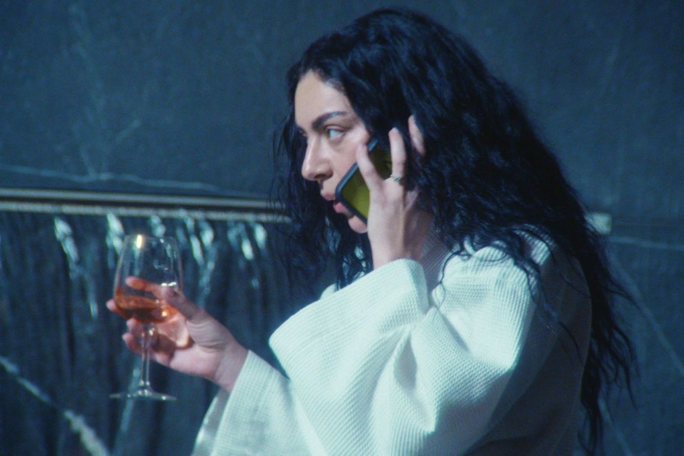 Szenenbild zum Film The Moment: Eine Person mit langen, dunklen, lockigen Haaren trägt einen hellen Bademantel und hält ein Glas mit bernsteinfarbenem Getränk in der rechten Hand. Der Hintergrund zeigt eine dunkle Wand mit einer goldfarbenen horizontalen Linie. [erstellt mit KI]