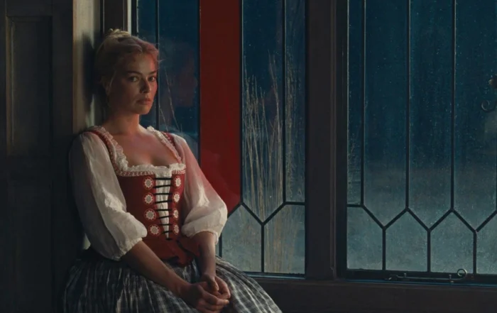 Eine Frau in einem Kleid mit rot-weißem Ober- und blaukarierten Unterteil sitzt auf einem Fenstersims. Draußen ist Nacht, während sie nicht zum Fenster sieht.