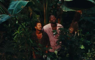 Szenenbild aus Taxi Moto: Zwei Personen stehen inmitten dichter, tropischer Vegetation. Große, grüne Blätter und verschiedene Pflanzen umgeben sie. Die Kleidung ist gemustert und in gedeckten Farben gehalten. Das Licht fällt durch das Blätterdach und erzeugt Schatten auf den Pflanzen und den Personen. [erstellt mit KI]