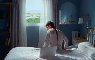 Szenenbild aus Stallion and a Crystal Ball: eine Person mit einem langen dünnen Zopf sitzt auf einem weißbezogenen Bett Richtung Fenster gerichtet, das Zimmer ist in dunklem blau gestrichen, es sind ein Regal und ein Spiegel zu sehen, durch das Zimmerfenster fällt Tageslicht