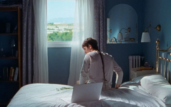 Szenenbild aus Stallion and a Crystal Ball: eine Person mit einem langen dünnen Zopf sitzt auf einem weißbezogenen Bett Richtung Fenster gerichtet, das Zimmer ist in dunklem blau gestrichen, es sind ein Regal und ein Spiegel zu sehen, durch das Zimmerfenster fällt Tageslicht