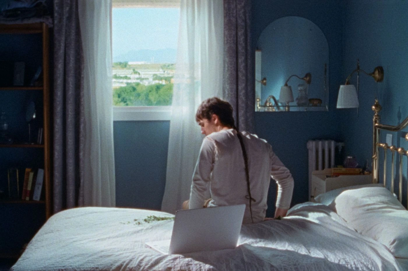 Szenenbild aus Stallion and a Crystal Ball: eine Person mit einem langen dünnen Zopf sitzt auf einem weißbezogenen Bett Richtung Fenster gerichtet, das Zimmer ist in dunklem blau gestrichen, es sind ein Regal und ein Spiegel zu sehen, durch das Zimmerfenster fällt Tageslicht