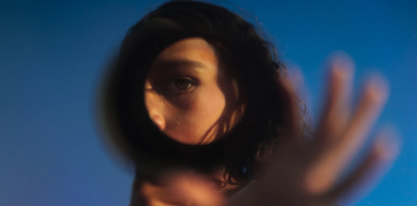 Eine Person blickt durch ein rundes Loch in einer blauen Fläche. Man sieht ein Auge und einen Teil des Gesichts. Eine Hand ist unscharf im Vordergrund erhoben. [erstellt mit KI]