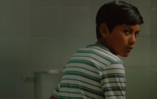 Szenenbild aus The River Train: Ein Kind im gestreiften T-Shirts sitzt mit dem Rücken zur Kamera und dreht seinen Kopf leicht zur Kamera.