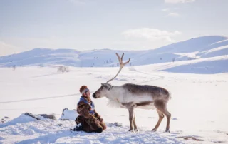 Szenenbild von Arru: Eine Frau im dicken Pelzmantel kniet im Schnee vor einer weiten Schneelandschaft vor einem Rentier.