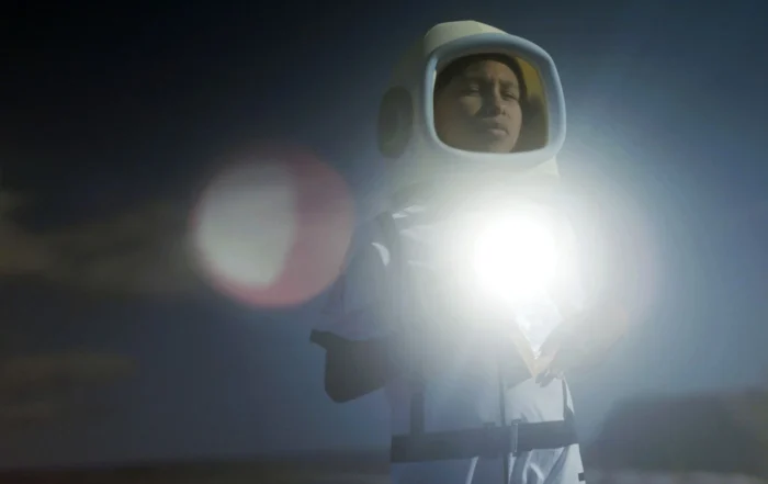 Eine Person trägt einen weißen Schutzanzug mit Kapuze und hält eine helle Lichtquelle vor dem Körper. Der Hintergrund ist dunkel und unscharf, mit Lichtreflexen und verschwommenen Formen. [erstellt mit KI]