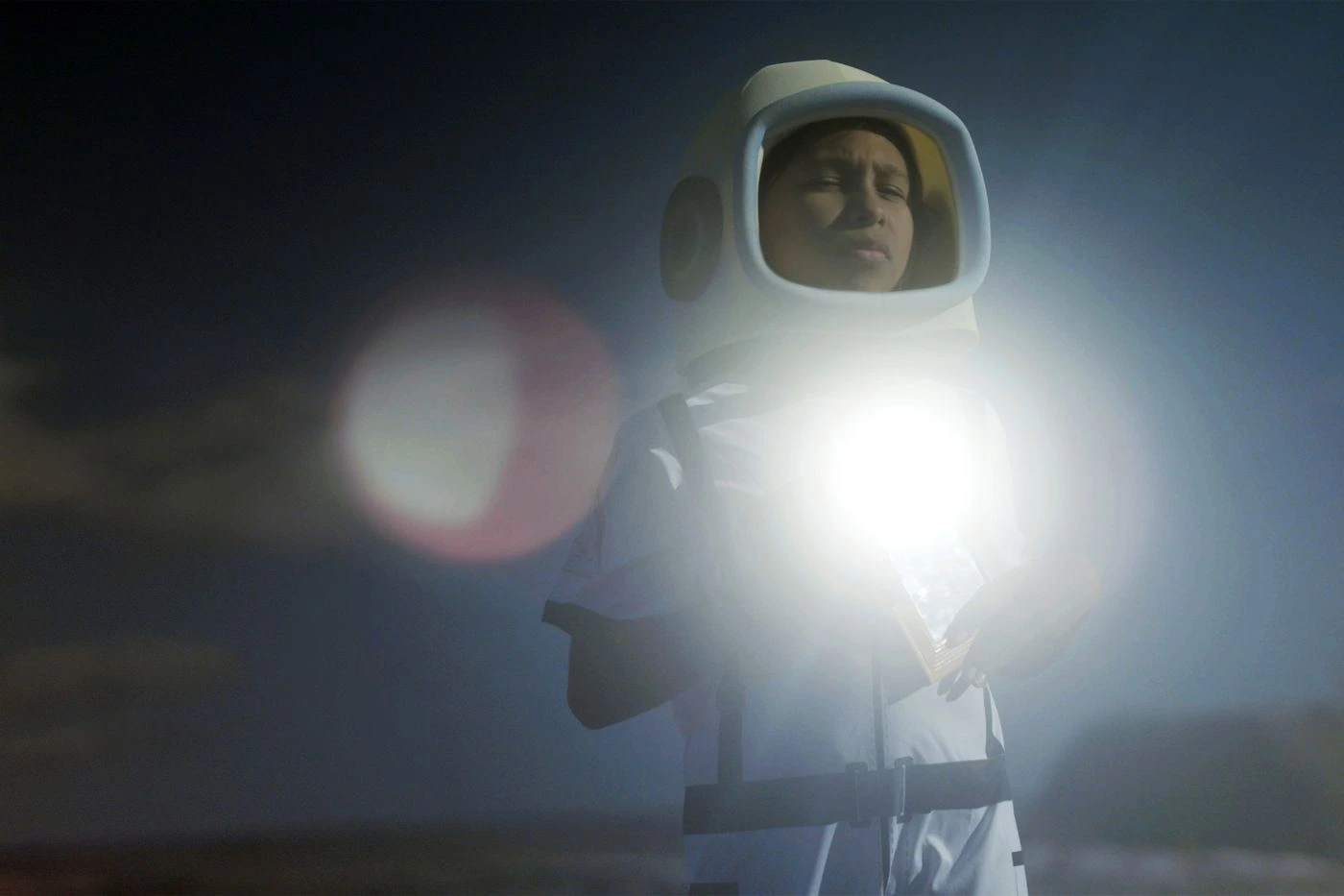 Eine Person trägt einen weißen Schutzanzug mit Kapuze und hält eine helle Lichtquelle vor dem Körper. Der Hintergrund ist dunkel und unscharf, mit Lichtreflexen und verschwommenen Formen. [erstellt mit KI]