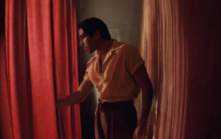 Szenenbild aus dem Film Narciso: Eine Person mit dunklem, zurückgekämmtem Haar trägt ein kurzärmeliges, helles Hemd und eine braune Hose. Sie steht zwischen zwei schweren, roten Vorhängen und hält einen der Vorhänge leicht geöffnet. Das Licht ist warm und gedämpft, wodurch eine intime Atmosphäre entsteht. [erstellt mit KI]